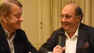 Two men sitting at a table, engaged in conversation, speaking into microphones.