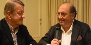 Two men sitting at a table, engaged in conversation, speaking into microphones.